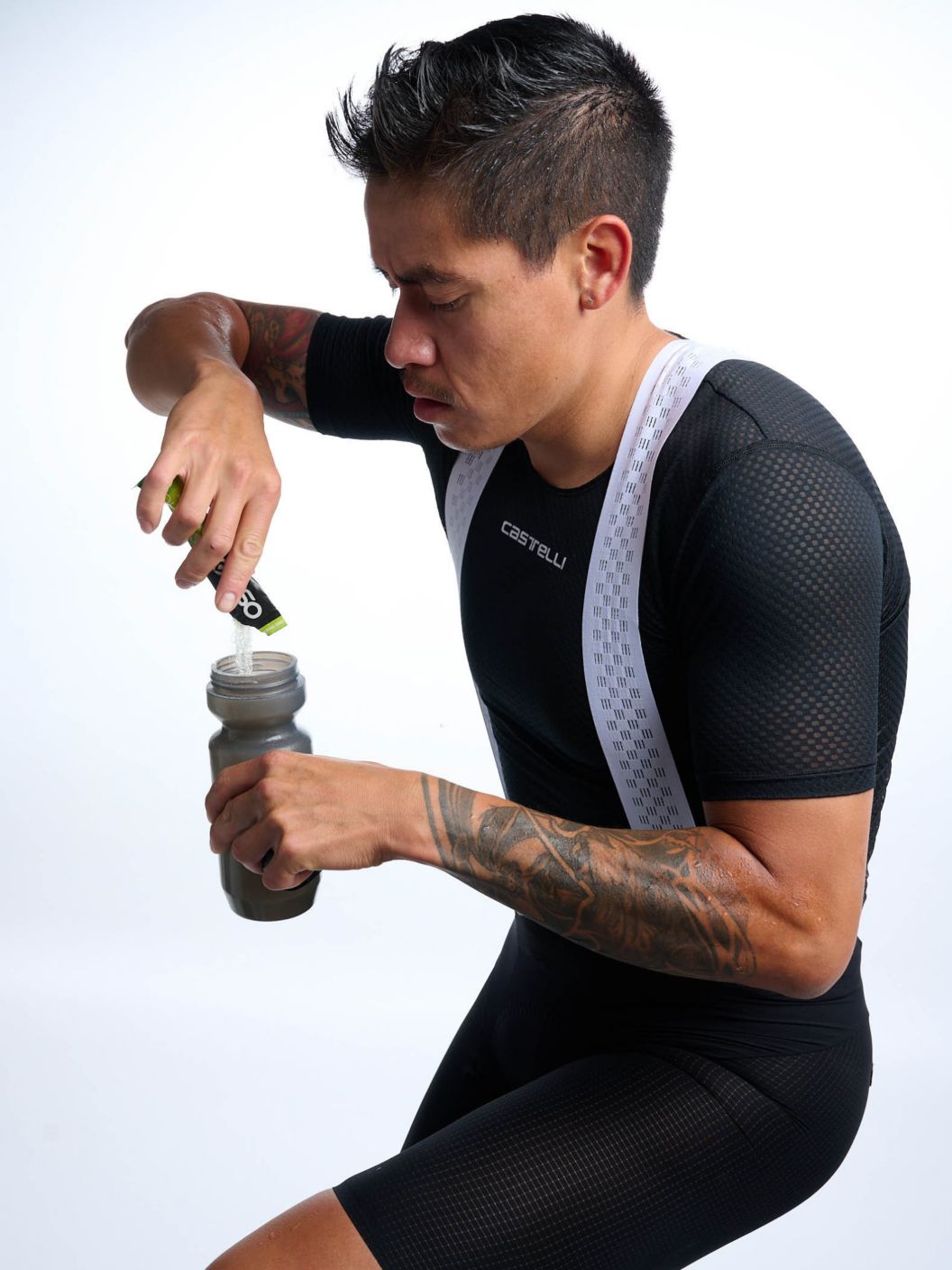A rider in a baselayer and bibs pours drink mix powder into a bottle. 